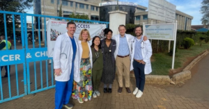 Doctors wearing white coats and scrubs pose with adults dresses in business professional attire in front of a hospital gate in Kenya.