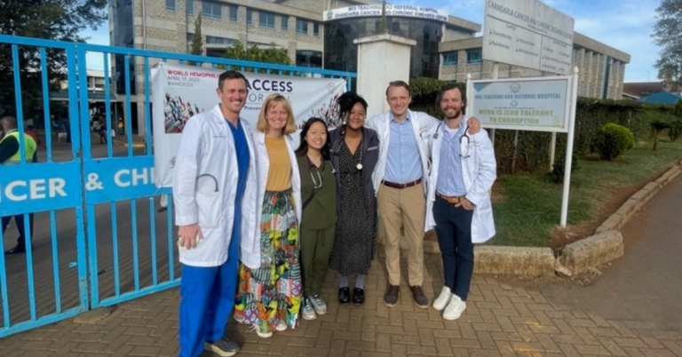 Doctors wearing white coats and scrubs pose with adults dresses in business professional attire in front of a hospital gate in Kenya.