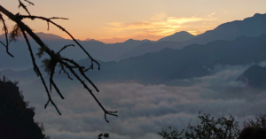 Sunset over dusty dark blue and grey mountains sit behind a bed of clouds.