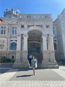 Female in blue dress and white tote bag poses in front of a large stone structure with columns and many windows.