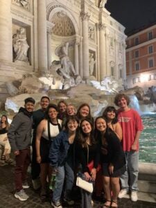 A group of friends smile while standing in front of the Trevi fountain in Italy.