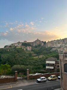 Mountains of Siena, Italy with stone churches and buildings behind a blue and yellow cloudy sky.