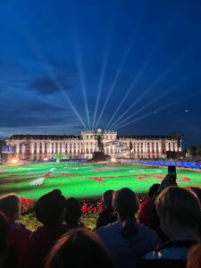 Outdoor concert at night in front of a large rectangular palace. A light garden contrasts the deep blue sky.