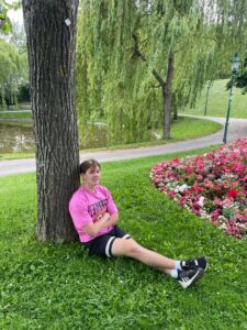 A student in a pink t-shirt and black shorts sits under a tree in a park.