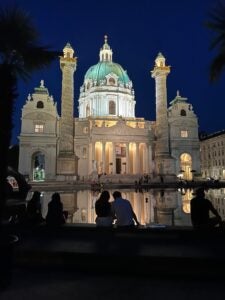 A Palace at night sits in front of a small body of water. The Palace has a green dome and symmetrical columns and windows.