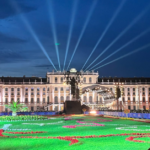 Outdoor concert at night in front of a large rectangular palace. A light garden contrasts the deep blue sky.