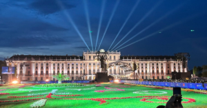 Outdoor concert at night in front of a large rectangular palace. A light garden contrasts the deep blue sky.