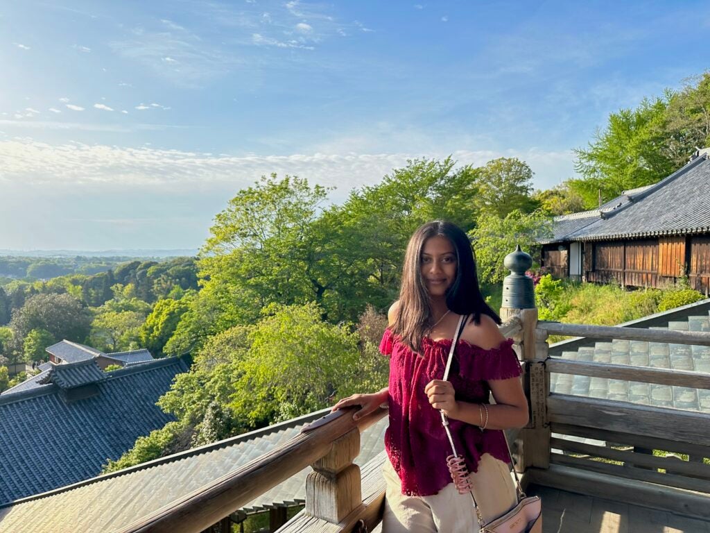A female in a red top and white pants smiles while one hand lays on the railing next to a large drop off.
