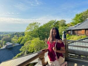 A female in a red top and white pants smiles while one hand lays on the railing next to a large drop off. 