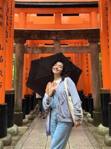 Girl with black umbrella, blue jeans, a grey sweater, and a canvas tote bag poses while visiting a Japanese shrine. The shrine is made of orange, black, and natural brown wooden posts with Japanese symbols inscribed onto each post.