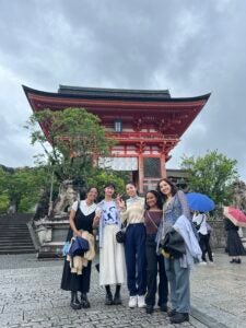 Four students dressed in casual clothing pose in front of a temple in Kyoto. The temple is red and has a traditional Japanese style roof.