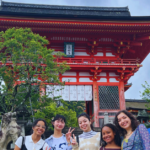 Four students dressed in casual clothing pose in front of a temple in Kyoto. The temple is red and has a traditional Japanese style roof.