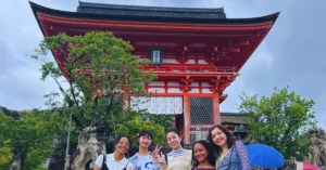 Four students dressed in casual clothing pose in front of a temple in Kyoto. The temple is red and has a traditional Japanese style roof.