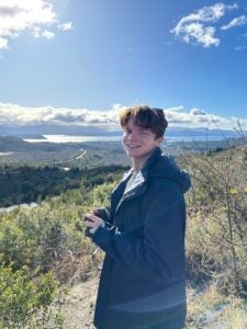 A student in a blue rain jacket poses for the camera amid bright blue skies and a valley of greenery behind him.