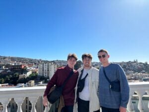 Three young men wear lightweight jackets and black nylon bags across their body as they smile for a photo. The background includes tall city buildings atop a hill