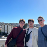 Three young men wear lightweight jackets and black nylon bags across their body as they smile for a photo. The background includes tall city buildings atop a hill
