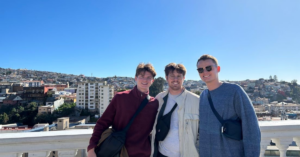 Three young men wear lightweight jackets and black nylon bags across their body as they smile for a photo. The background includes tall city buildings atop a hill