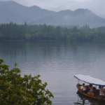 A small, handmade boat floats on top of still water. Dark grey mountains stand tall in the background behind a cloudy sky.