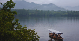 A small, handmade boat floats on top of still water. Dark grey mountains stand tall in the background behind a cloudy sky.
