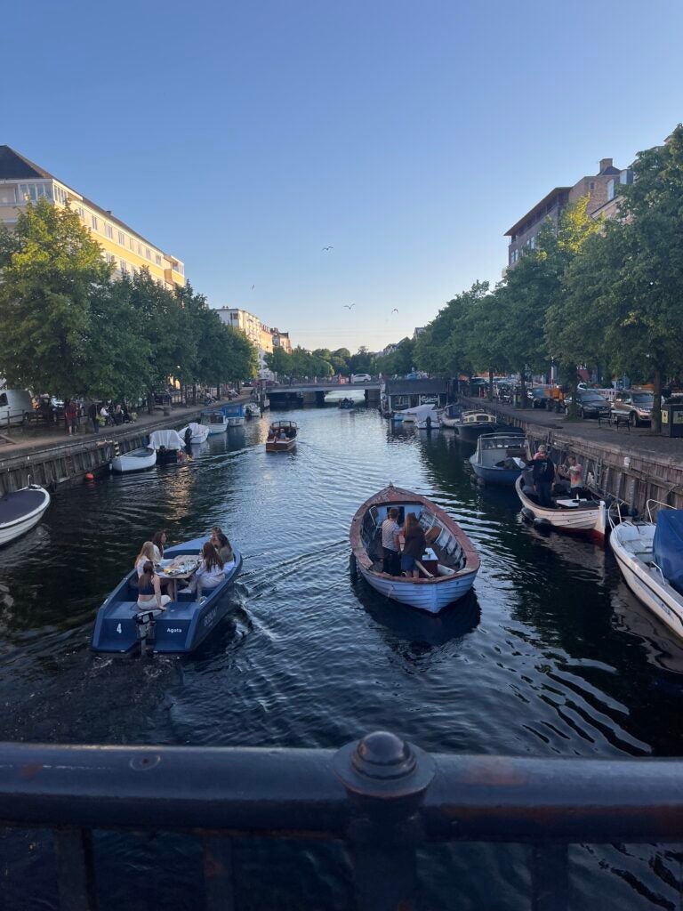 A canal with many row boats is captured during the daytime as the sun is peaking through the tall buildings that surround the canal. Trees also line the canal of each side.