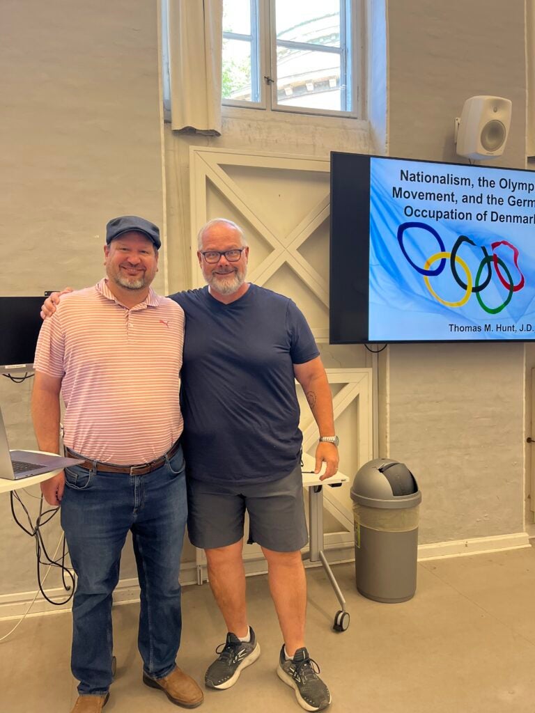 Two professors embrace and smile in front of a powerpoint. One professors wears blue jeans and an orange shirt while the other wears grey shorts and a blue tee.