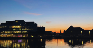 Still water during sunset is a dark, deep blue. The sky is turning from blue to cream to slightly pink as the sun sets behind an illuminating building and small house that sits on the waterfront.