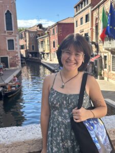 A student with short brown hair, bangs, glasses and a black shoulder bag smiles while on a bridge in Venice
