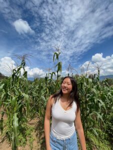 Girl in white top and blue shorts smiles with her head slightly bend towards one side. Tall stalks of vegetation are behind her as she poses.