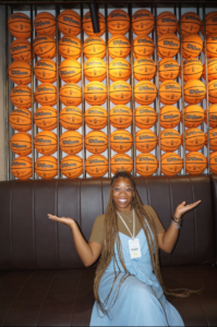 A young lady in a green top and blue overalls poses in front of a wall of basketballs lined up towards the sky.