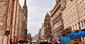 An old, European town is the focal point as the street is bustling with locals crossing the streets, cars crowding the intersections and outdoor seating being enjoyed by all.