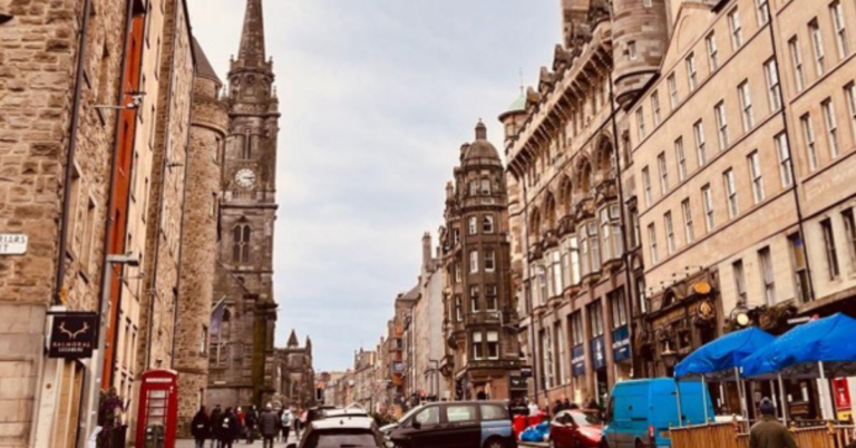 An old, European town is the focal point as the street is bustling with locals crossing the streets, cars crowding the intersections and outdoor seating being enjoyed by all.