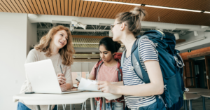 Three young women gather together around a table and discuss plans while filling out forms with large backpacks on their back.