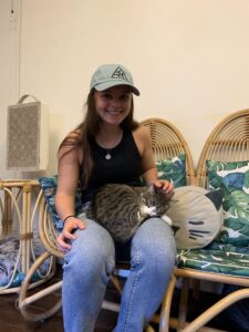 A female student with a green adidas hat, black top and blue jeans smiles as she holds a cat in her lap.