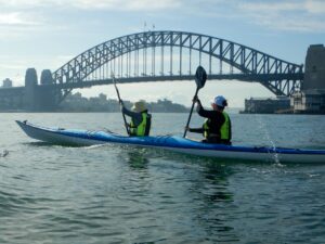 Two kayakers in mid-row in the Sydney Harbour look to be enjoying themselves. A large iron bridge is in the background. 