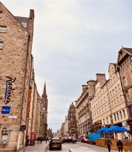 An old, European town is the focal point as the street is bustling with locals crossing the streets, cars crowding the intersections and outdoor seating being enjoyed by all.