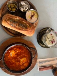 A small loaf of brea, hummus, olives, a light spread and a tomato paste soup sits on top of a table next to a glass of green iced matcha.