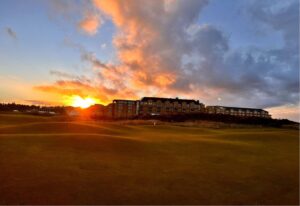 The sun is setting behind a large building in front of a green golf course.