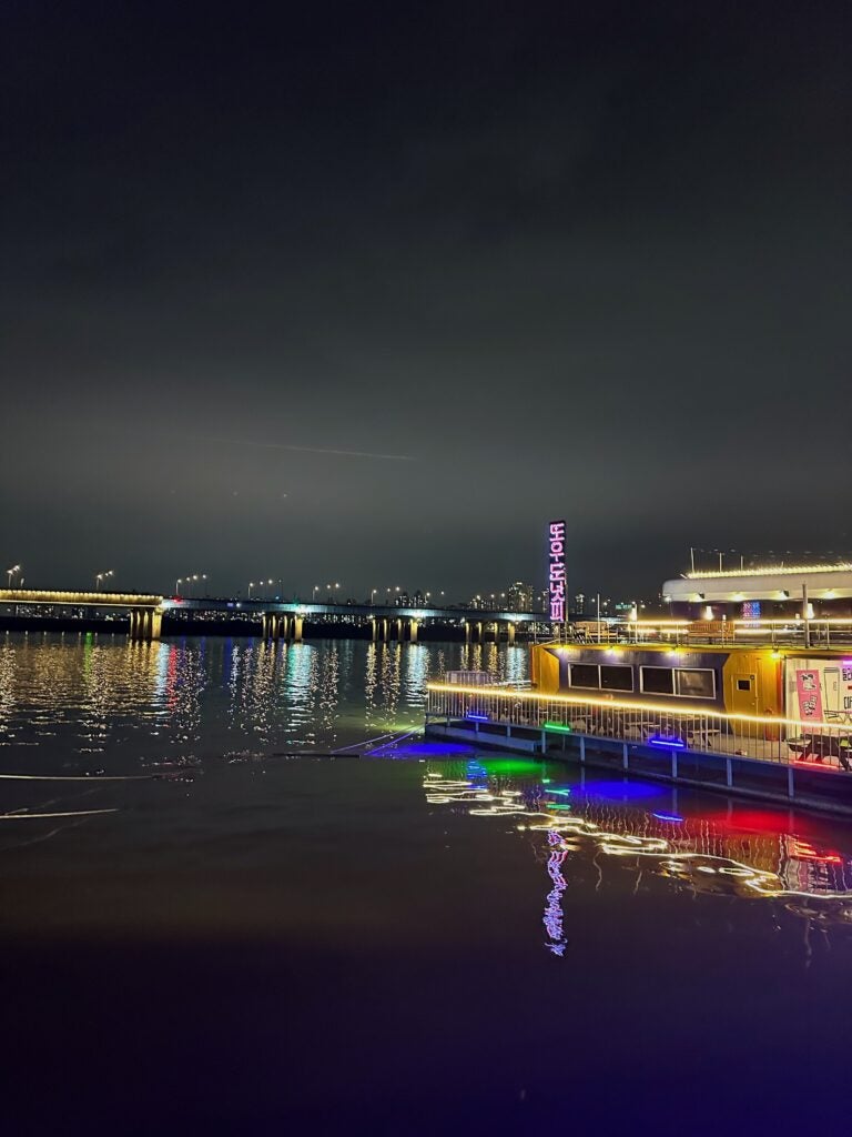 A dock is lit at night with red, green, and blue river lights. There are buildings in the background and a clear, dark grey sky overhead.