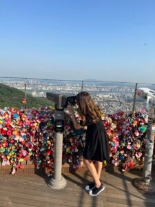 A student in a black dress and brown hair looks through a telescope to overlook a large city.