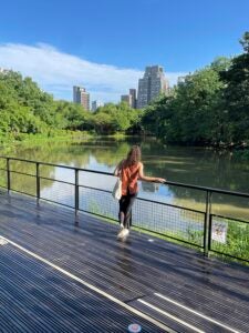 A woman stands in a park in front of a pond with many trees and a city skyline in the background.