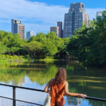 A woman stands in a park in front of a pond with many trees and a city skyline in the background.