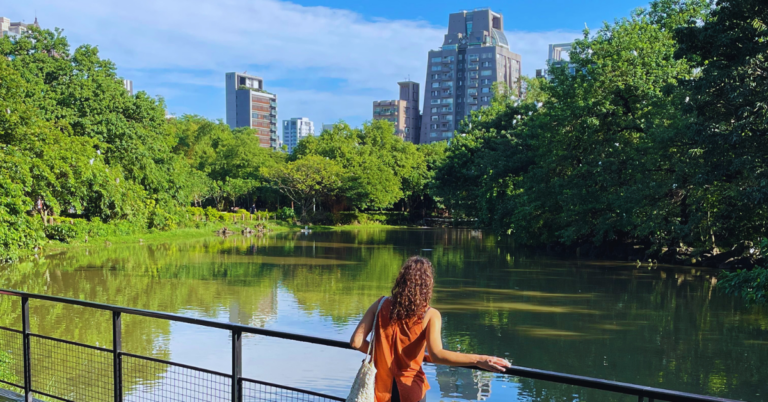 A woman stands in a park in front of a pond with many trees and a city skyline in the background.
