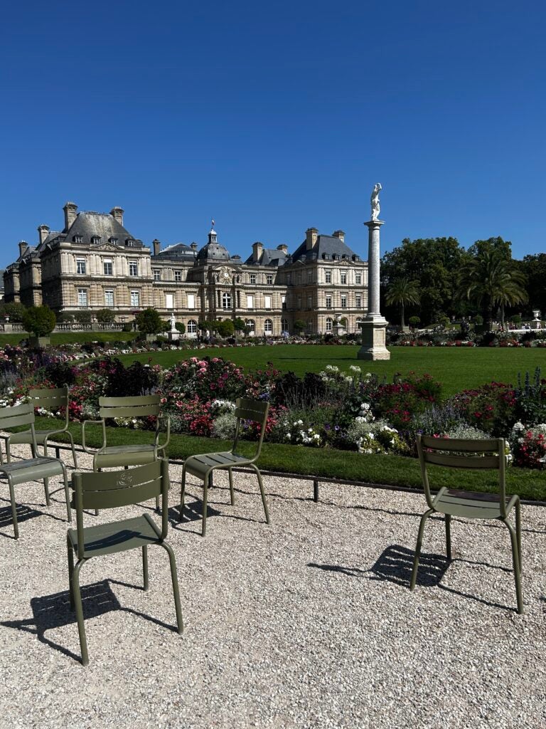 An old, charming building in Paris with a beautiful garden in front. A pebble walkway with iron picnic tables and chairs leads through the garden, adding to the rustic charm of the scene.