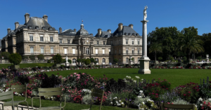 An old, charming building in Paris with a beautiful garden in front. A pebble walkway with iron picnic tables and chairs leads through the garden, adding to the rustic charm of the scene.