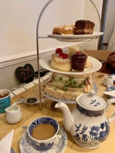 A traditional British afternoon tea set on a table. It includes a tiered tray with assorted finger sandwiches, scones with clotted cream and jam, and an array of delicate pastries. A teapot and teacup with saucer are placed alongside the treats.