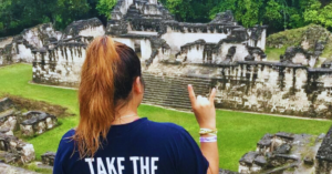 A student wearing a blue UT t-shirt kneels while overlooking ancient ruins.