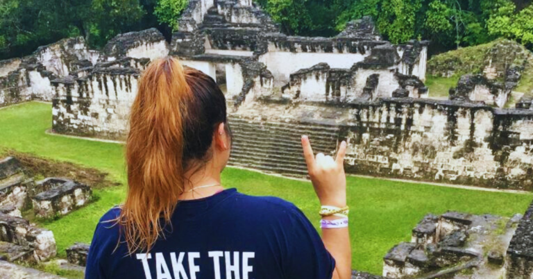 A student wearing a blue UT t-shirt kneels while overlooking ancient ruins.