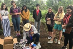 Students stand around a man kneeling on the floor combing through archaeological items.