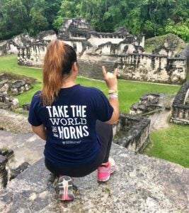 A student wearing a blue UT t-shirt kneels while overlooking ancient ruins.