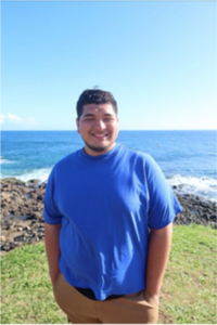 A male student in s blue tee shirt stands near a cliff on a grassy area with the sea behind him smiling on a sunny day.